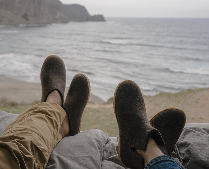 Two people relaxing with their feet up facing the ocean, wearing glerups slip-ons in Chestnut, brown and boots in Chestnut, brown, both with honey rubber soles.