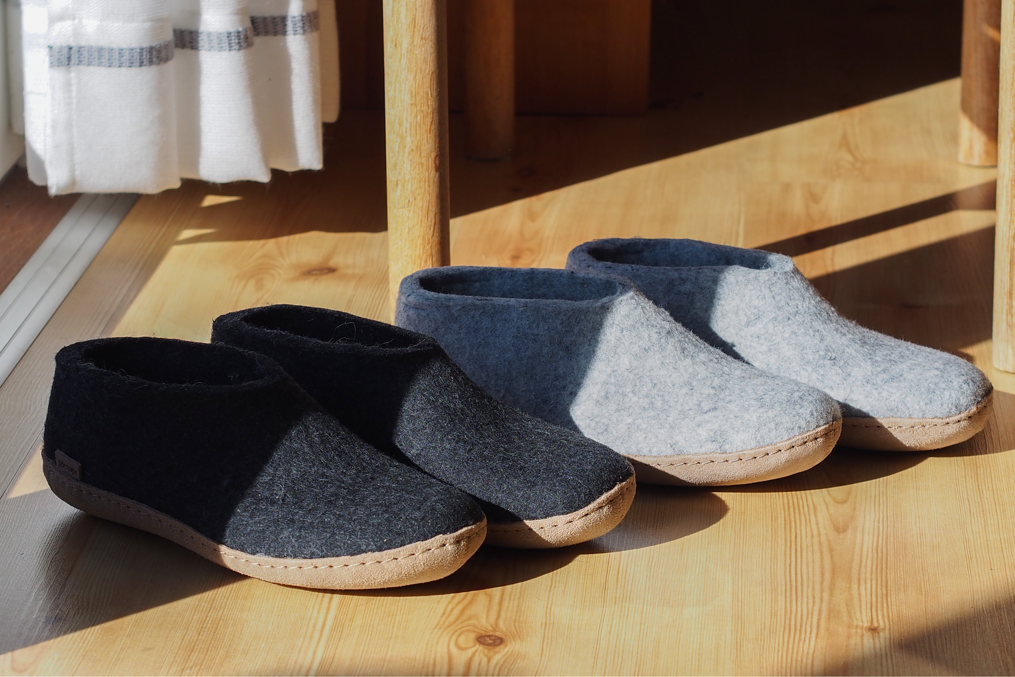 An indoor, high-angle shot of two pairs of glerups shoes sitting in a patch of bright morning sunlight on a light-colored hardwood floor near a wooden chair and a white striped curtain; one pair is charcoal, black and the other is Grey, and both have tan leather soles.